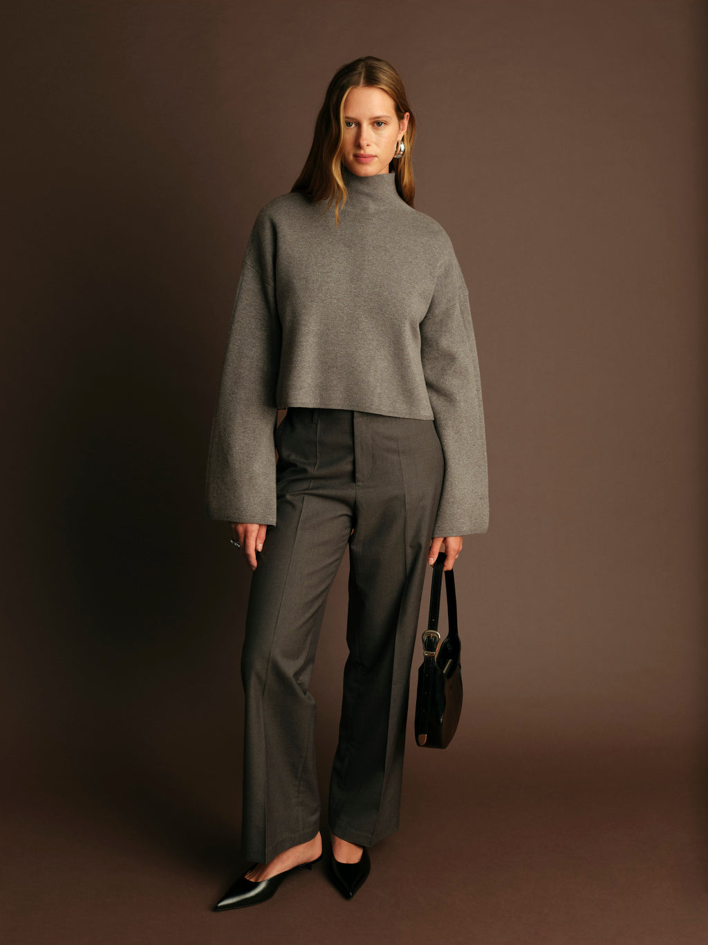 Woman wearing a gray turtleneck sweater and pants holding a black handbag against a brown background