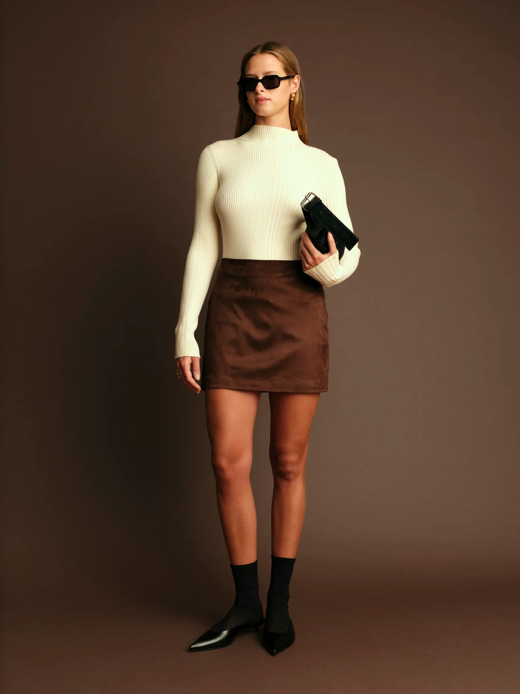Woman wearing a white turtleneck, brown skirt, and black shoes against a brown background