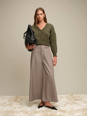 Woman wearing a green sweater and beige pants holding a black bag against a neutral background