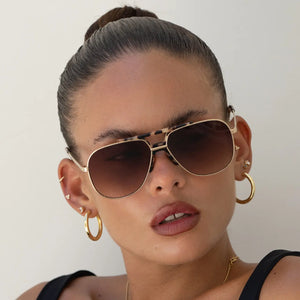 Woman wearing gold aviator sunglasses and hoop earrings against a neutral background