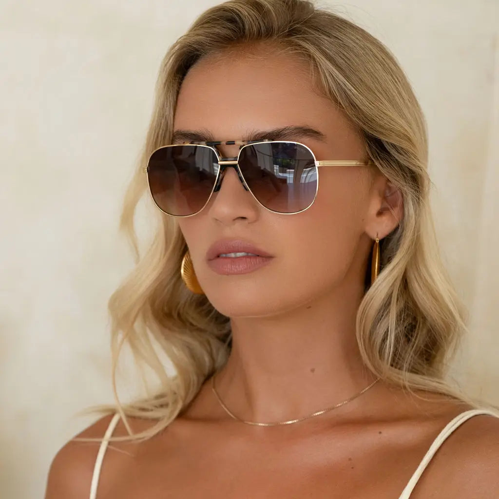 Woman wearing gold aviator sunglasses and hoop earrings with a neutral background