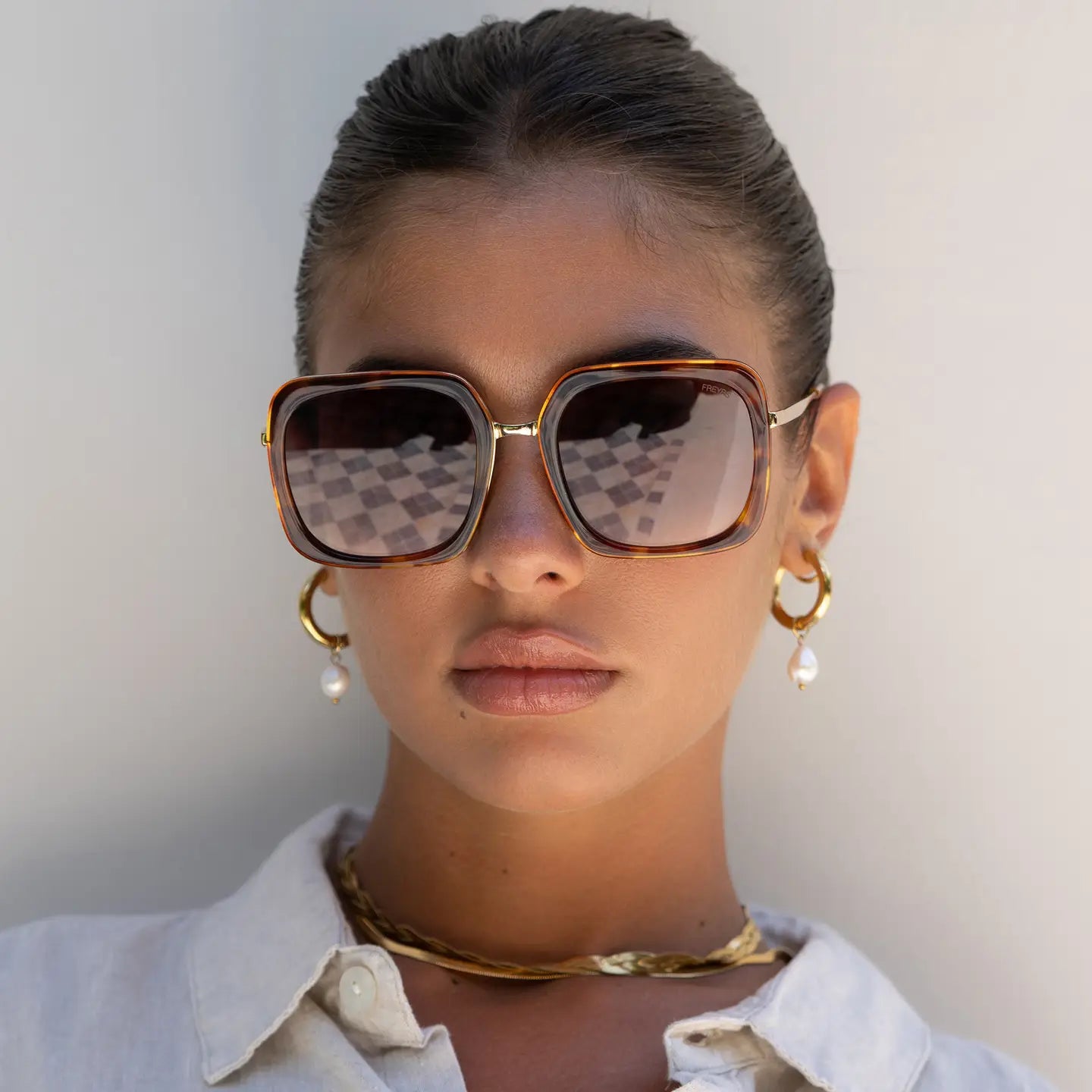 Woman wearing large sunglasses and gold jewelry against a neutral background