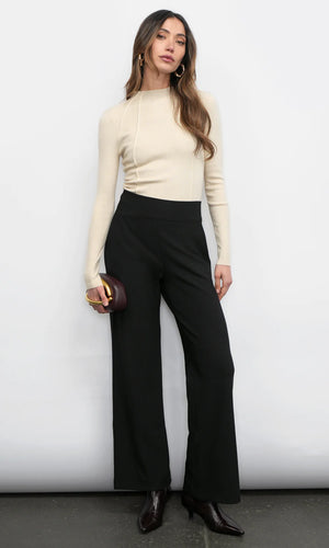 Woman wearing a beige long-sleeve top and black pants against a white background