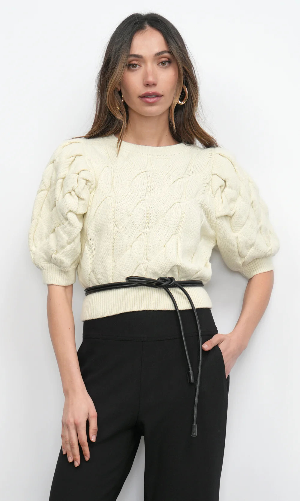 Woman wearing a cream knitted top and black pants against a white background