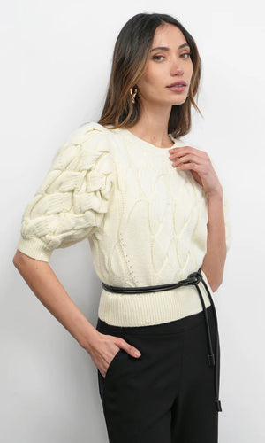 Woman wearing a cream cable knit sweater with black pants against a white background