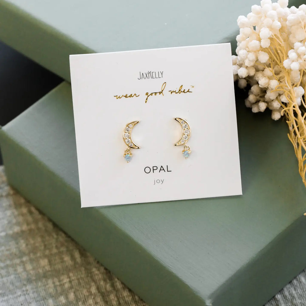 Gold moon-shaped earrings on a Jax Kelley branded card with a green box and decorative elements in the background.