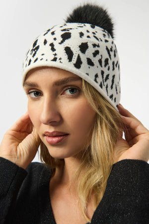Woman wearing a white beanie with black leopard print pattern and black pom-pom on a light gray background