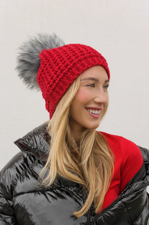 Woman wearing a red knit beanie with a gray pom-pom, black puffer jacket, and red shirt against a light background.