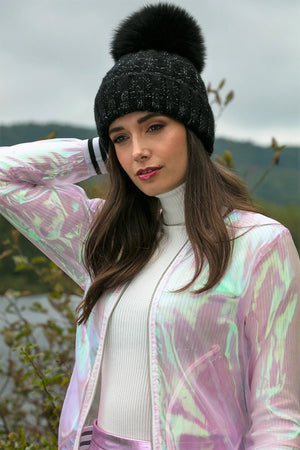 Woman wearing a black knit beanie with a pom-pom, standing outdoors with mountains in the background.