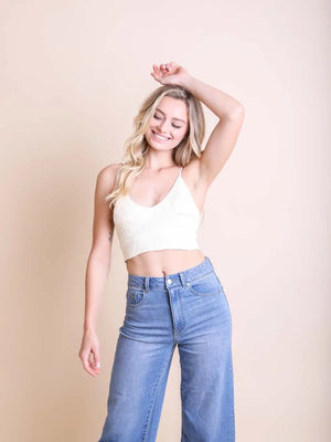 A woman wearing a white v-neck tank top and blue jeans, posing with one hand raised to her head.