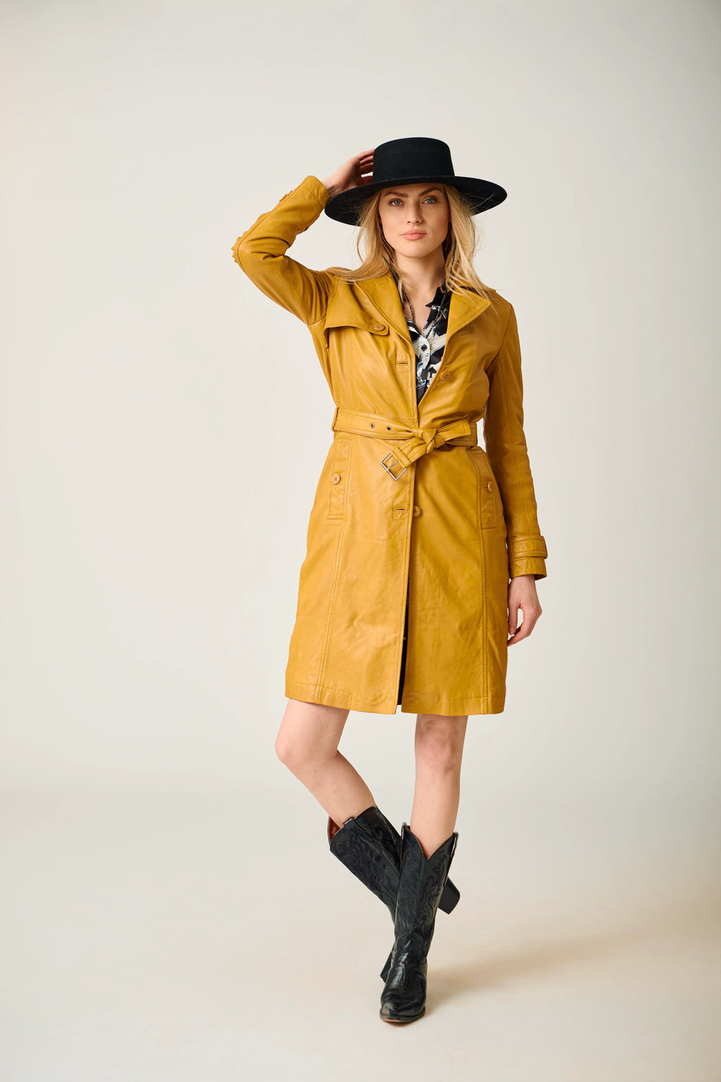Woman wearing a yellow trench coat, black hat, and black boots on a plain background