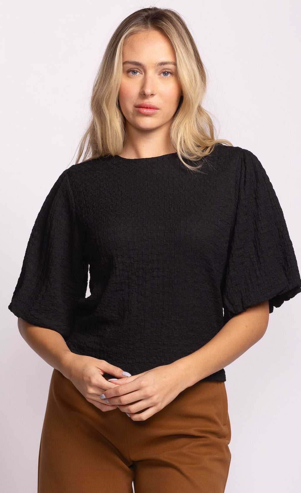 Woman wearing a black top and brown pants against a light pink background