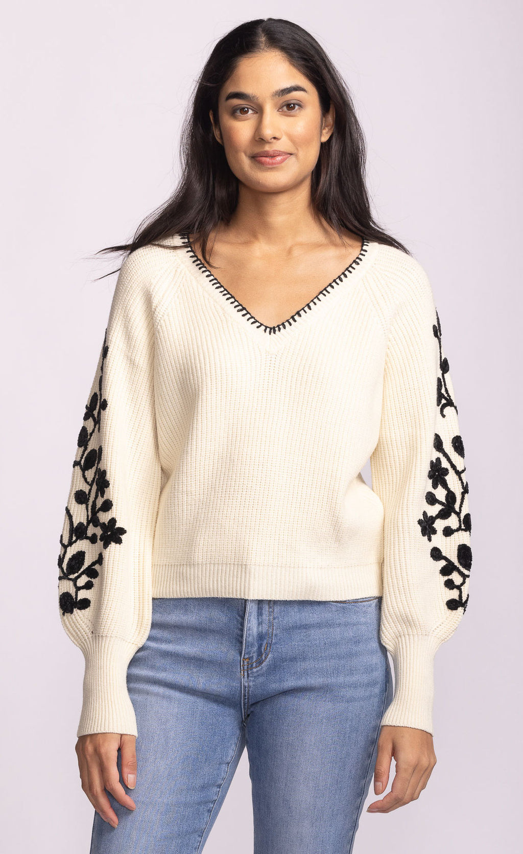 Woman wearing a cream sweater with black floral patterns on sleeves against a light purple background