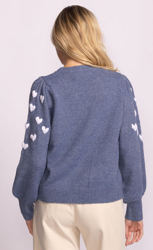 Blue sweater with heart patterns on sleeves worn by a person against a light pink background