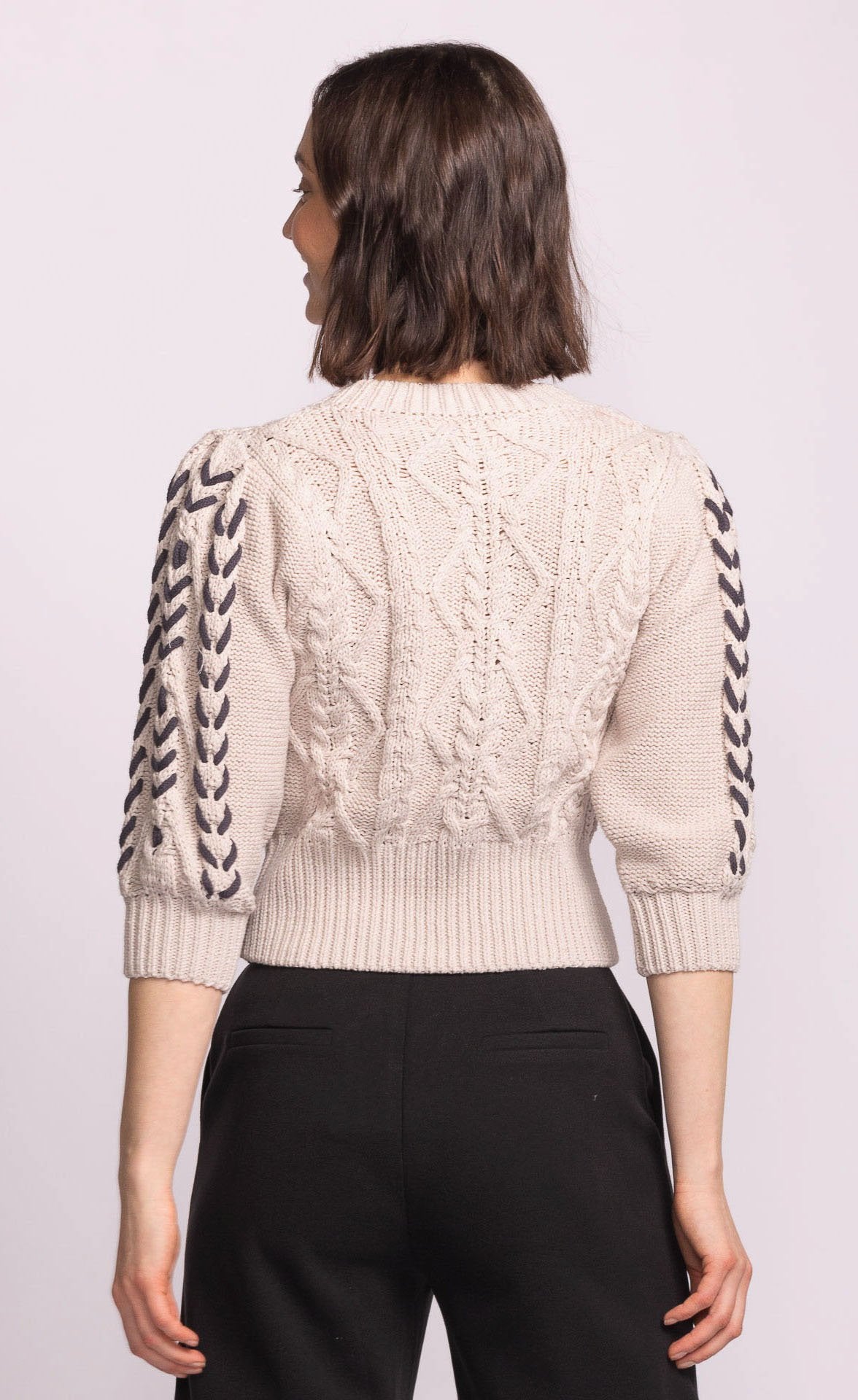 Person wearing a cream cable knit sweater with black braided details on sleeves against a light purple background
