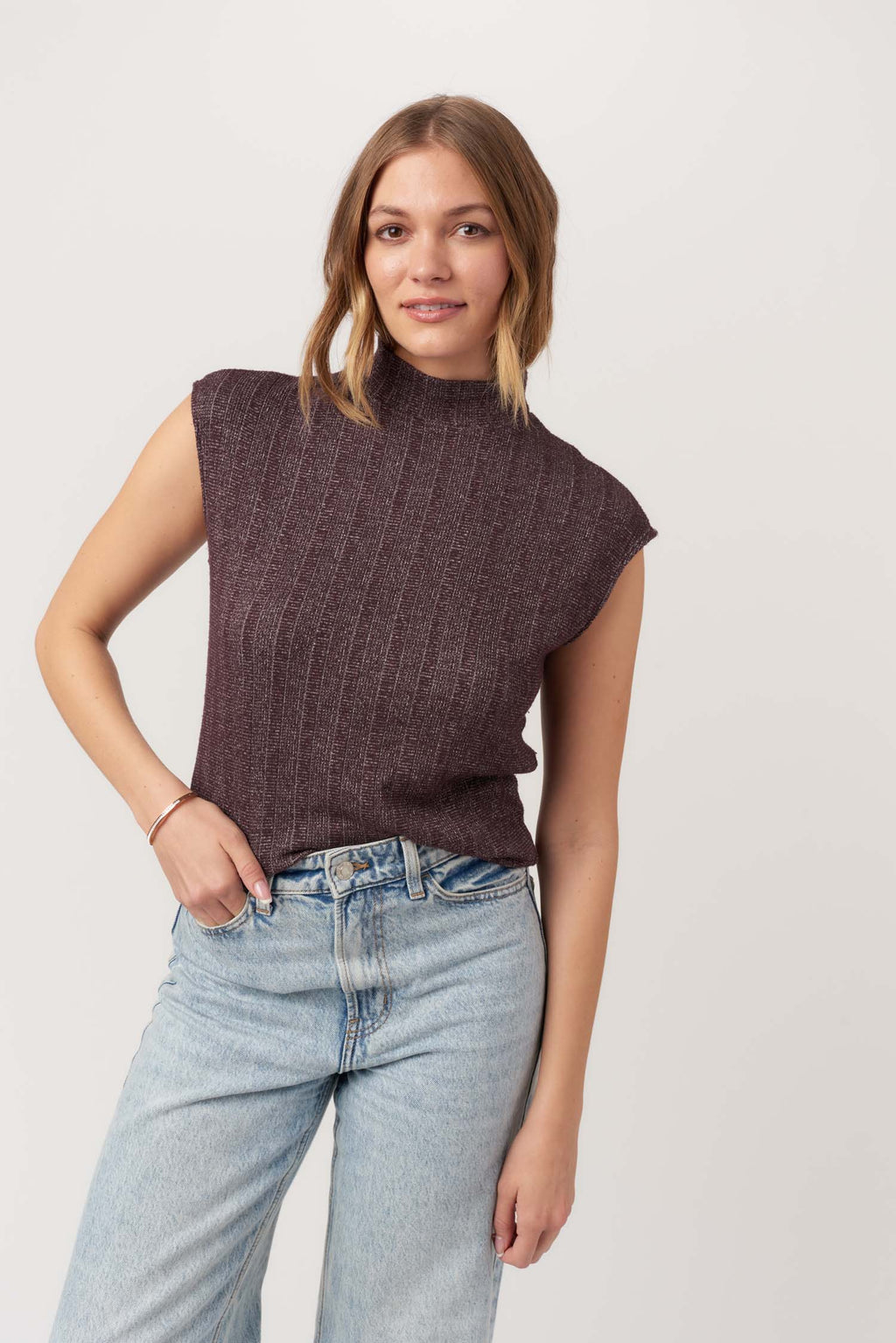 Woman wearing a brown sleeveless sweater and light blue jeans on a white background
