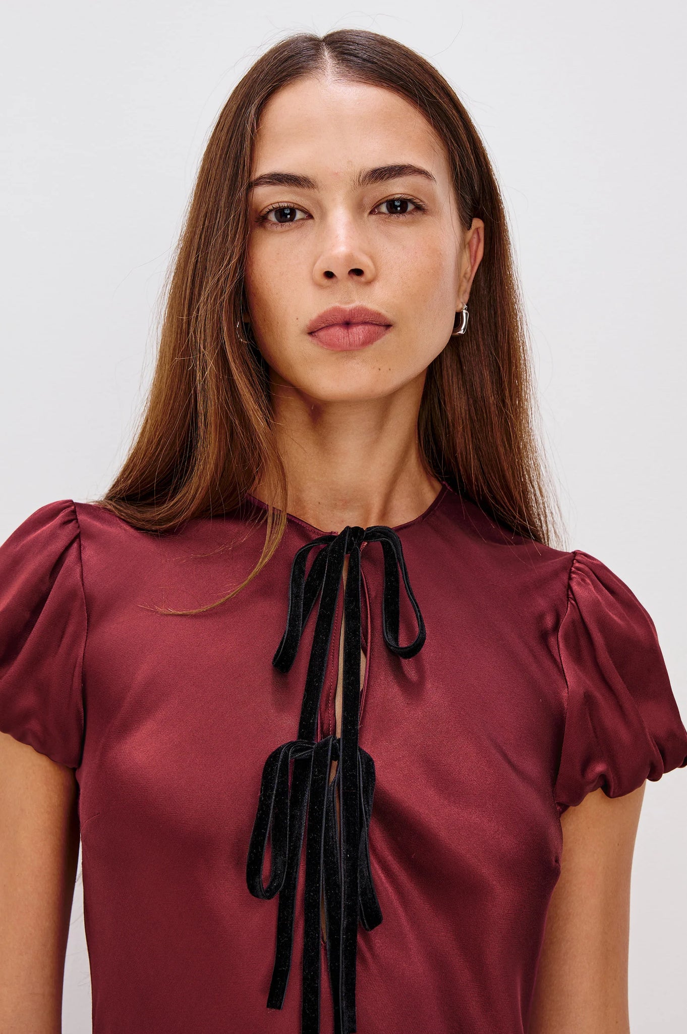 Woman wearing a burgundy blouse with black tie details against a plain background