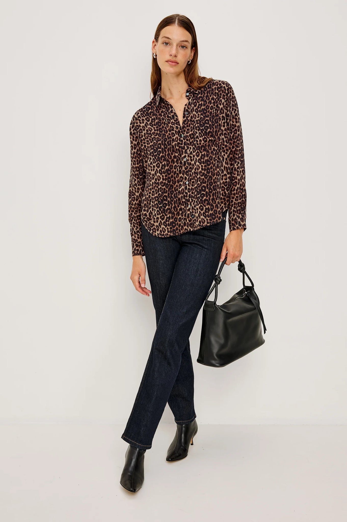 Woman wearing a leopard print blouse, dark jeans, and black boots holding a black handbag on a white background