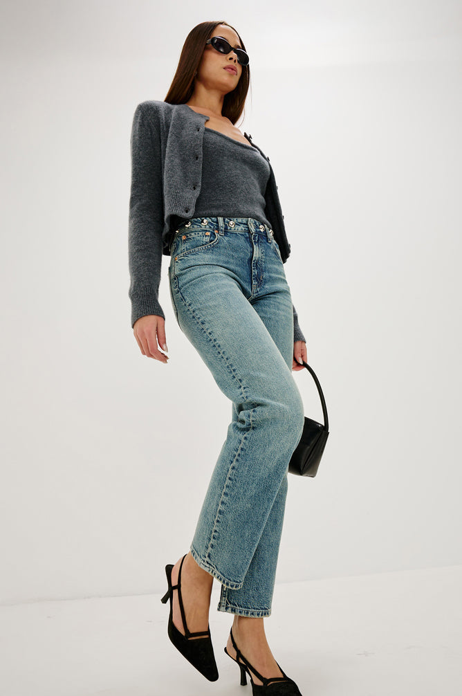 Woman wearing a gray cardigan, black top, and blue jeans on a white background