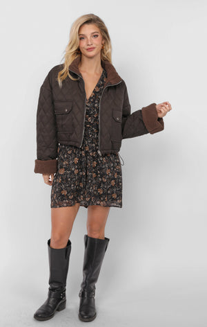 Woman wearing a brown jacket, floral dress, and black knee-high boots on a white background