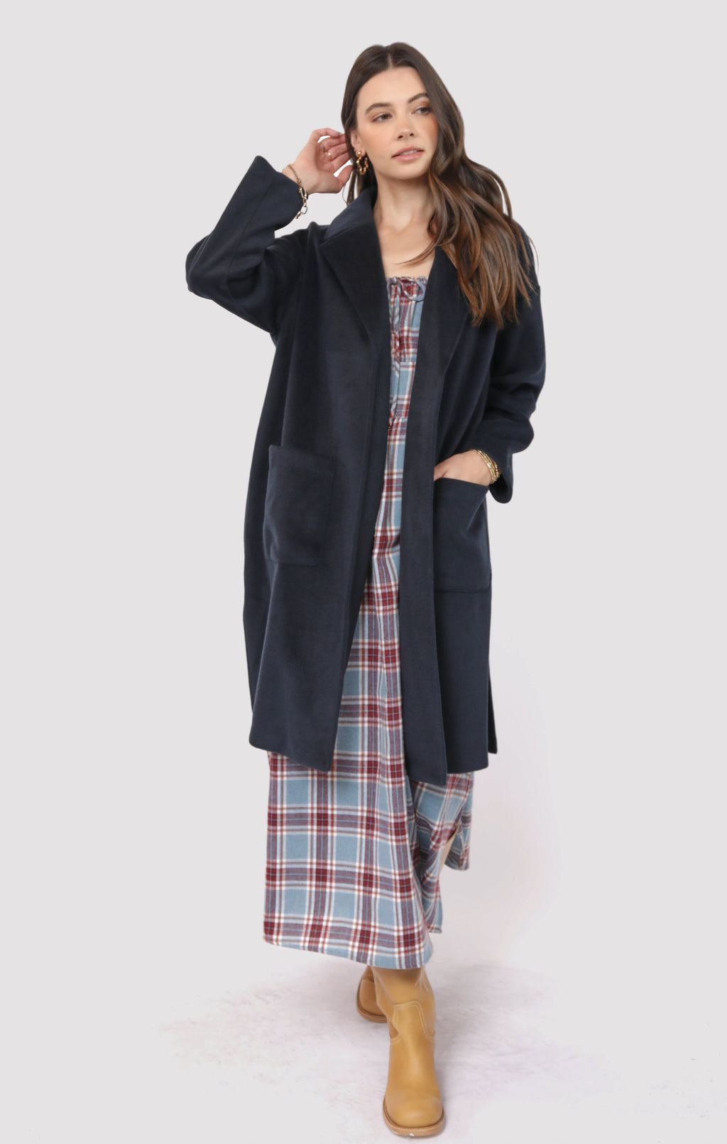 Woman wearing a long dark coat over a plaid dress with brown boots on a white background