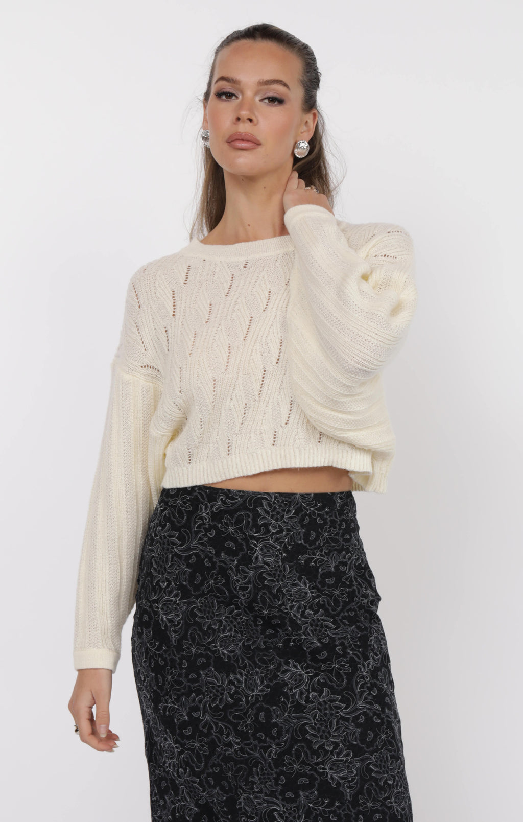 Woman wearing a cream cable knit sweater and black patterned skirt on a white background