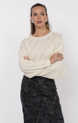 Woman wearing a cream cable knit sweater and black patterned skirt on a white background
