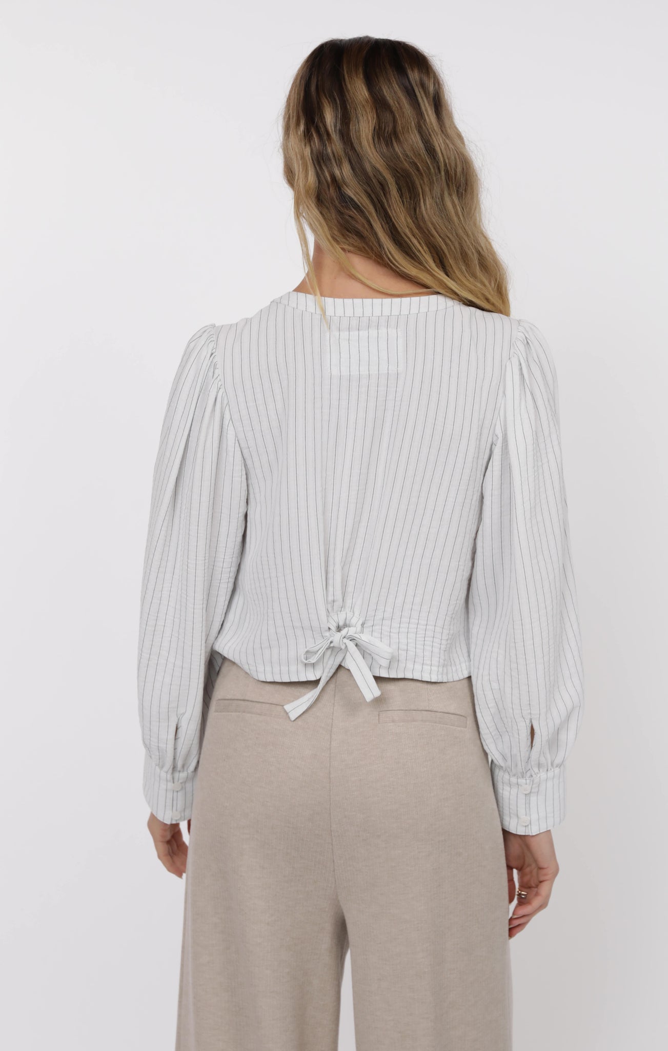 Woman wearing a light-colored striped blouse with a tie detail at the back, standing against a plain background.