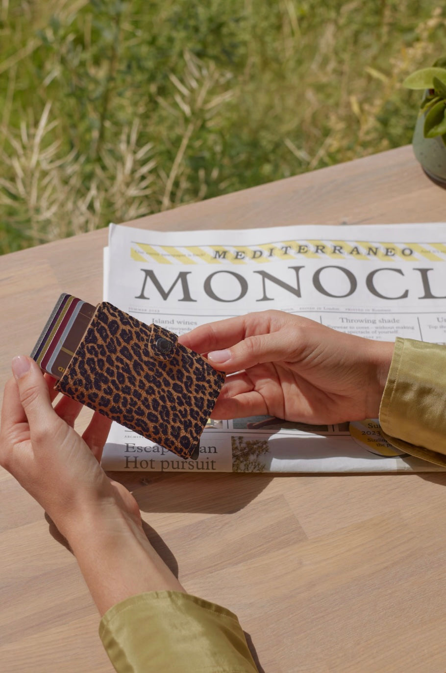 Person holding a leopard print wallet over a newspaper on a wooden table with a blurred natural background
