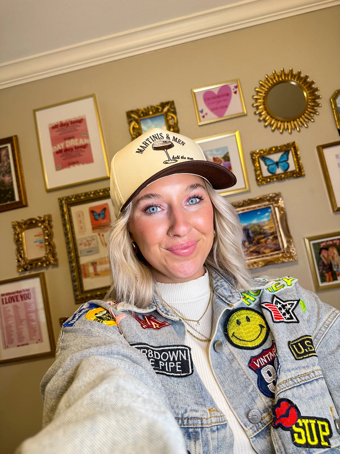 Person wearing a cap and patch-covered jacket in a room with framed pictures on the wall.