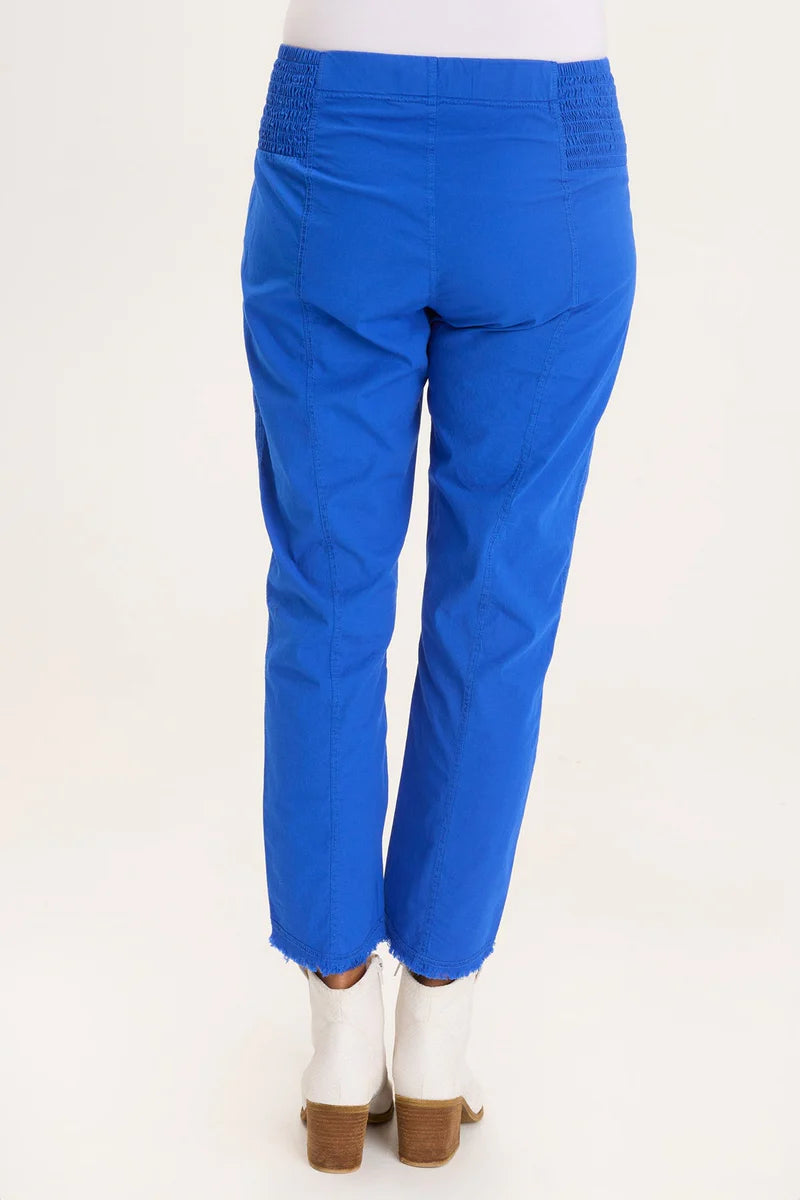 An ankle length slim fit pant with a pull on waist that features shirred panels, two inseam pockets, two patch zippered pockets, and a raw edge rounded hem.