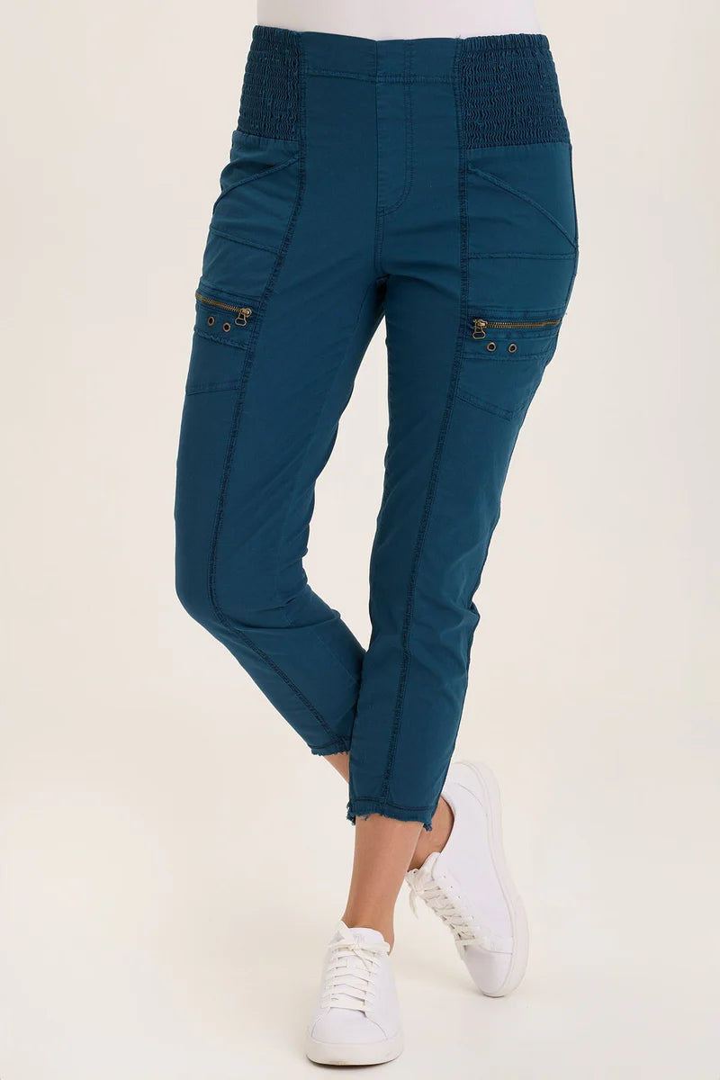 An ankle length slim fit pant with a pull on waist that features shirred panels, two inseam pockets, two patch zippered pockets, and a raw edge rounded hem.