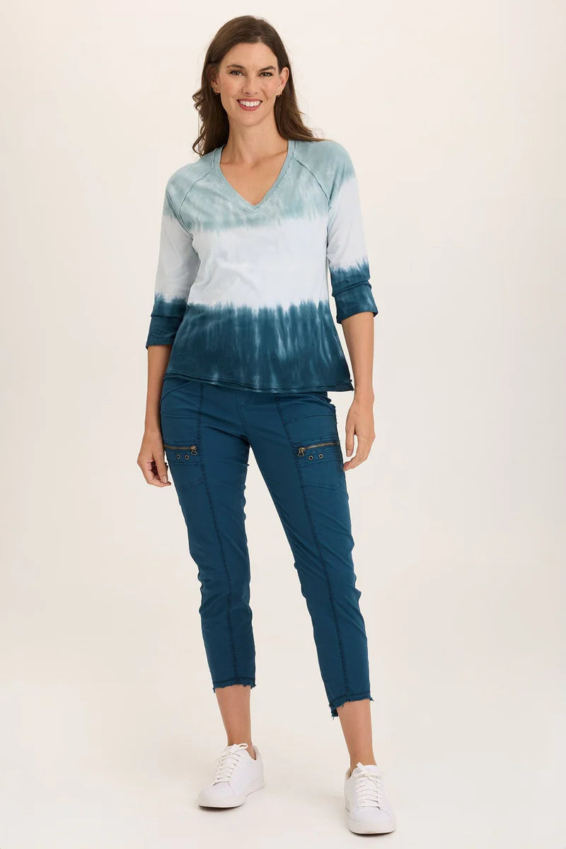 An ankle length slim fit pant with a pull on waist that features shirred panels, two inseam pockets, two patch zippered pockets, and a raw edge rounded hem.