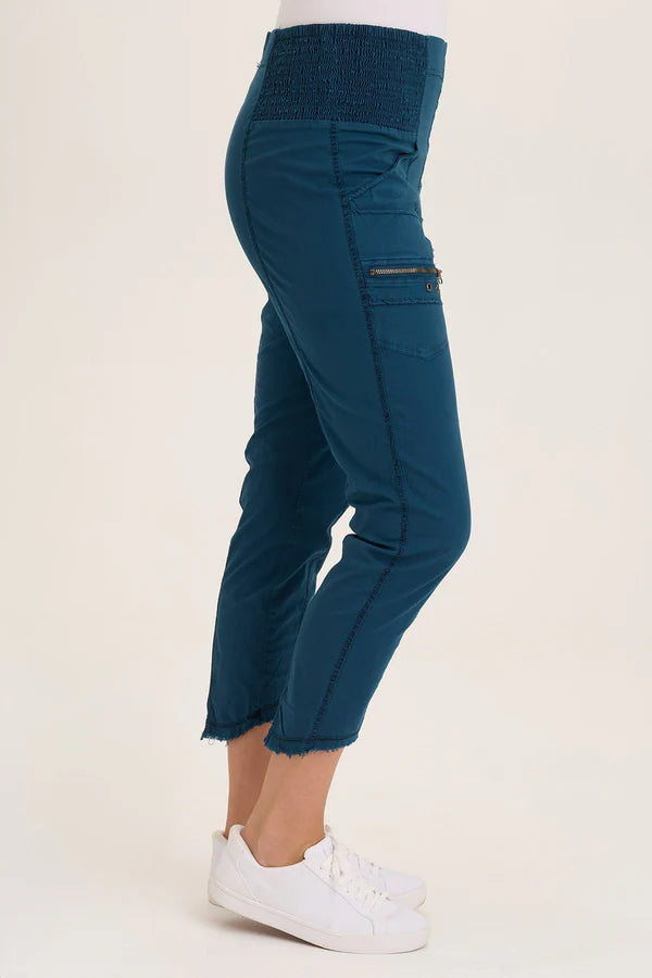 An ankle length slim fit pant with a pull on waist that features shirred panels, two inseam pockets, two patch zippered pockets, and a raw edge rounded hem.
