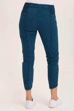 An ankle length slim fit pant with a pull on waist that features shirred panels, two inseam pockets, two patch zippered pockets, and a raw edge rounded hem.