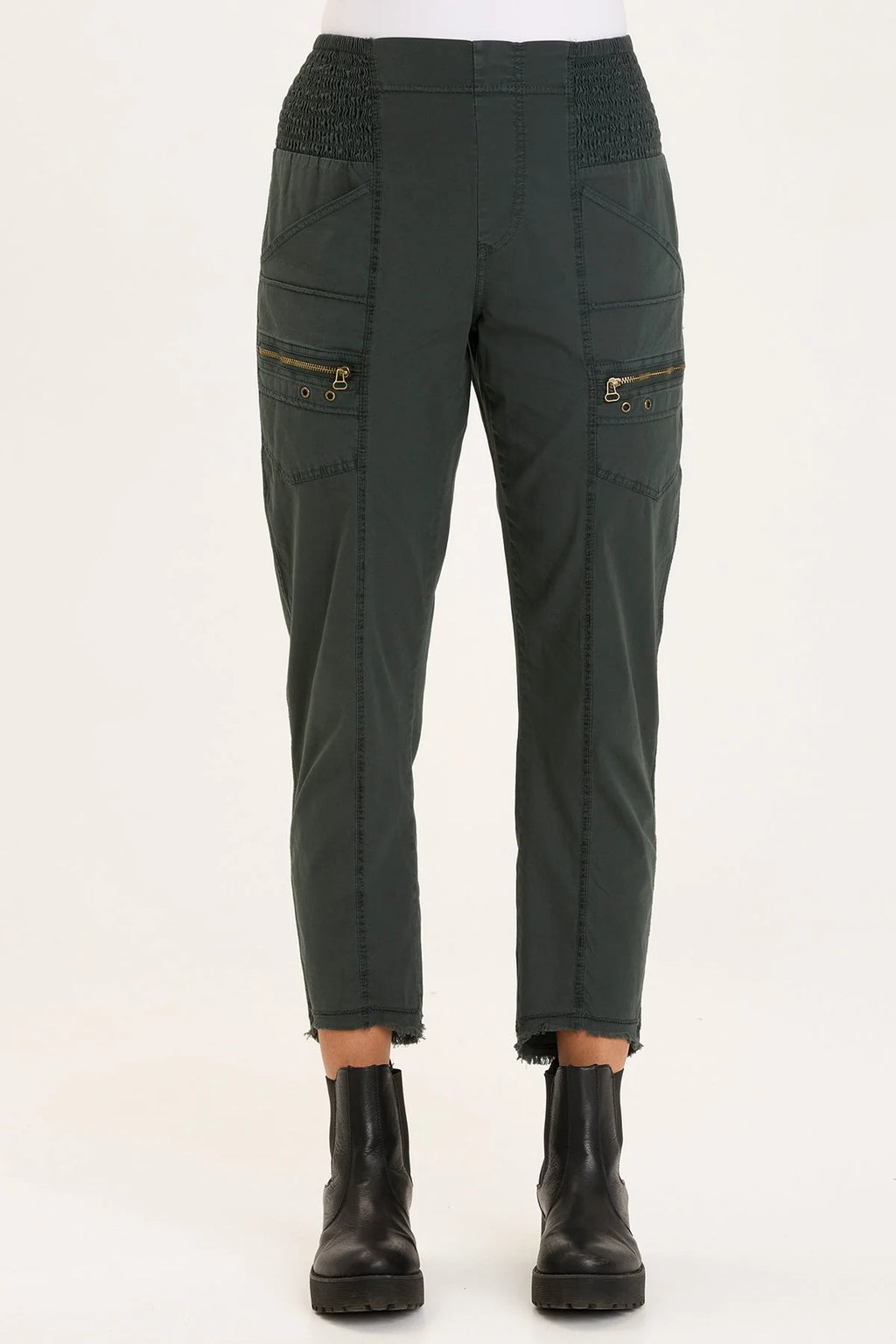 An ankle length slim fit pant with a pull on waist that features shirred panels, two inseam pockets, two patch zippered pockets, and a raw edge rounded hem.