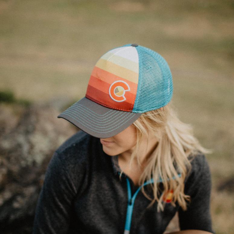 A person wearing a graphic-printed hat with a red to yellow gradient and a blue mesh, featuring a gray bill with blue contrast stitching and a green under-bill.